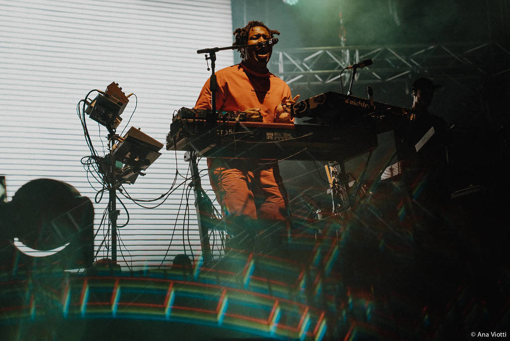 sampha performing onstage behind multiple pads and keyboards, in an orange jumpsuit