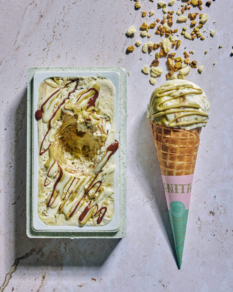 a tub of white chocolate and pistachio ice cream next to a cone with a scoop of the ice cream, both are drizzled with a green sauce and nuts
