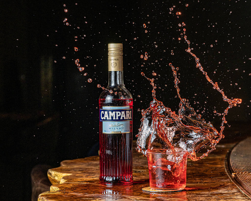 a bottle of campari next to a negroni that is mid-splash everywhere