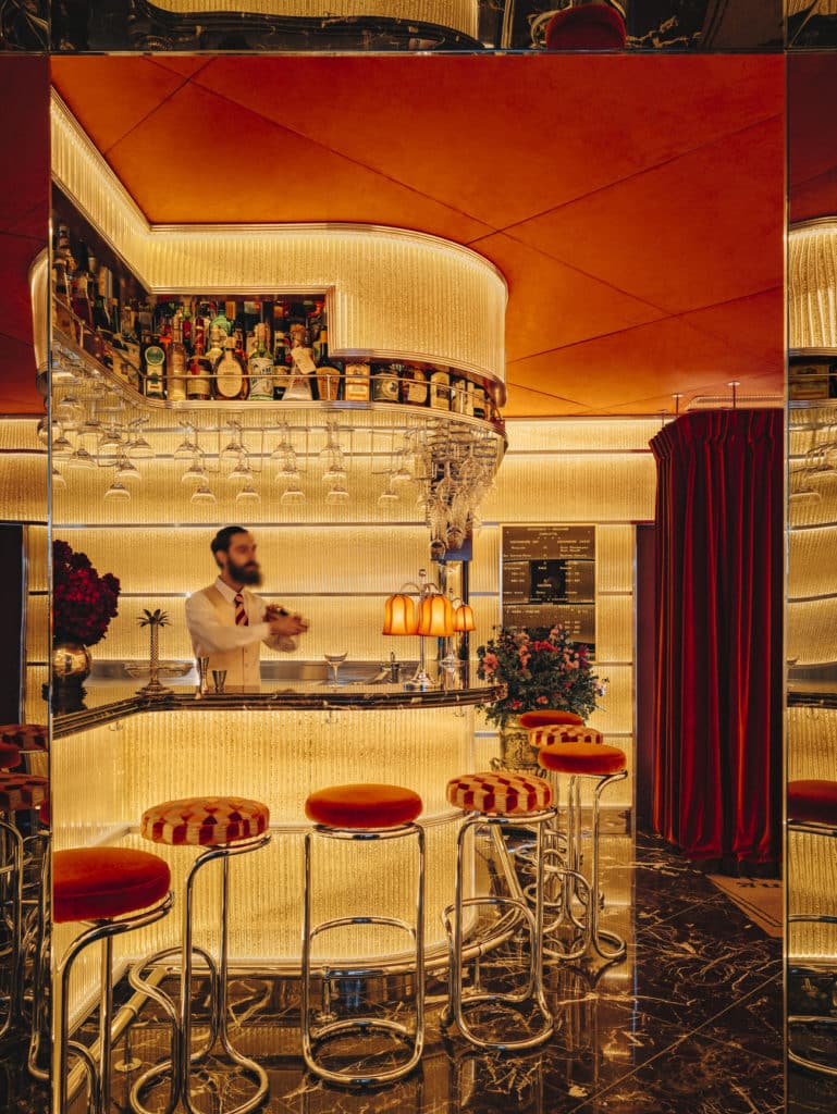 a bartender behind a glamorous, old-school looking bar with plenty of subdued yellow lighting, red barstools, and other red accents