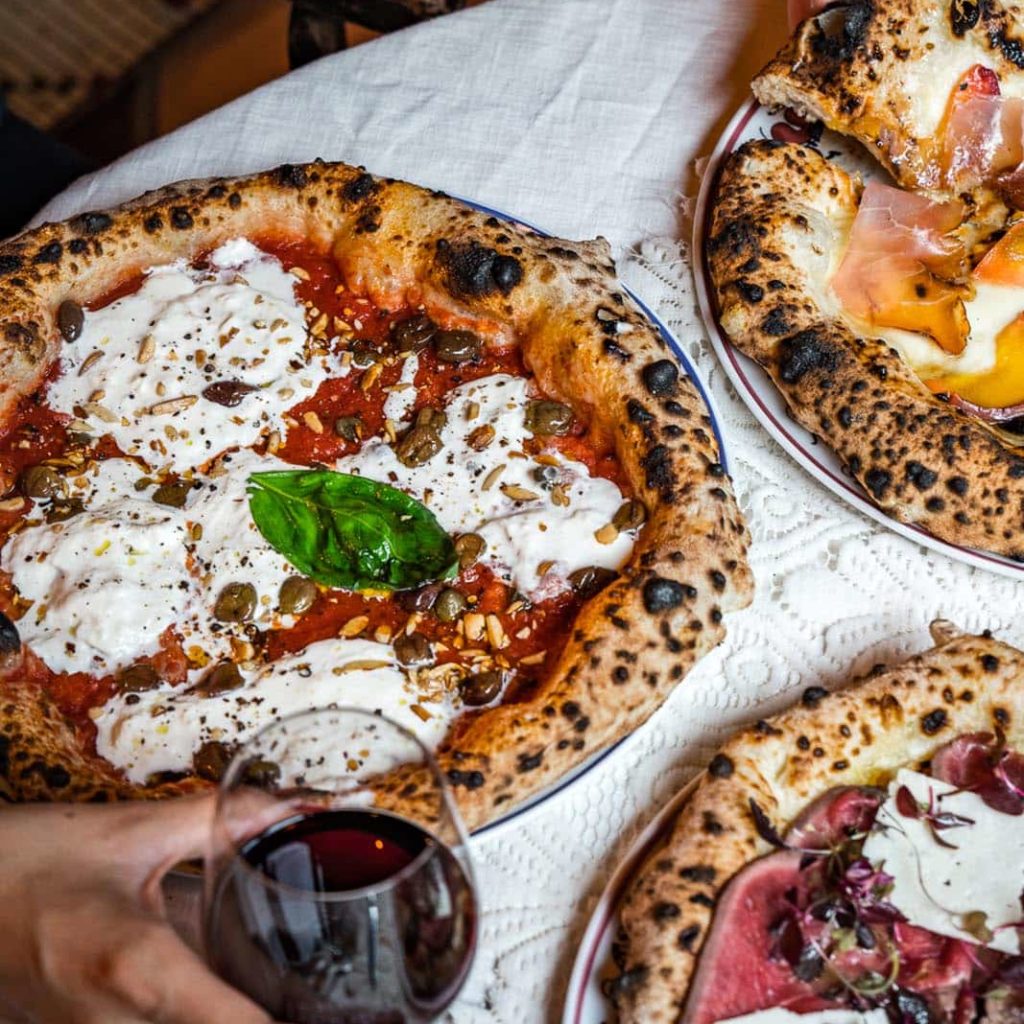 three pizzas spread on a table, with a hand holding a glass of red wine visible at the bottom of the image