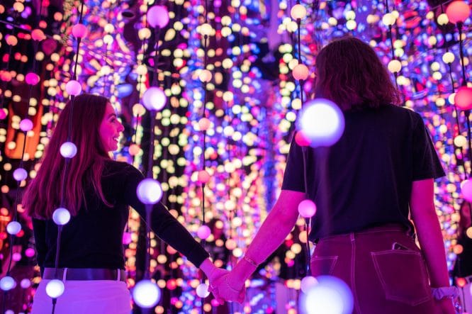 Two friends holding hands in a lit up installation at Dopamine Land