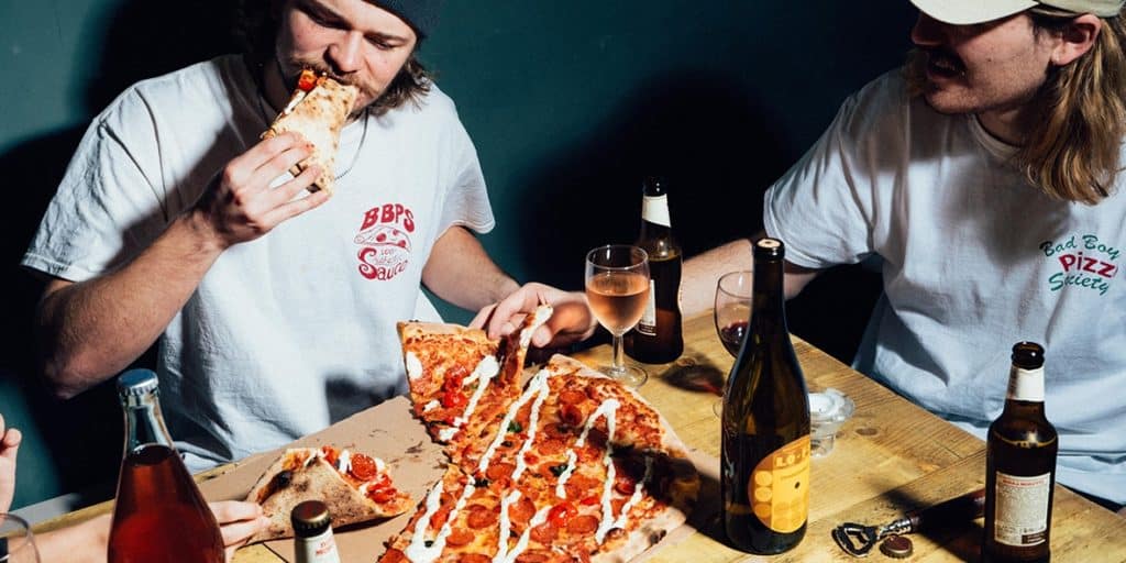 two people tucking into pizza slices, with bottles of wine and beer also visible on the table