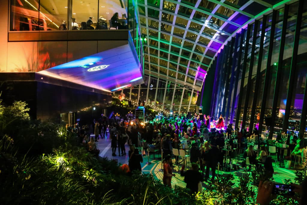 A photo of the Sky Garden Halloween Party, a sea of people dancing in the lowlit room with the open expansive view of London next to them