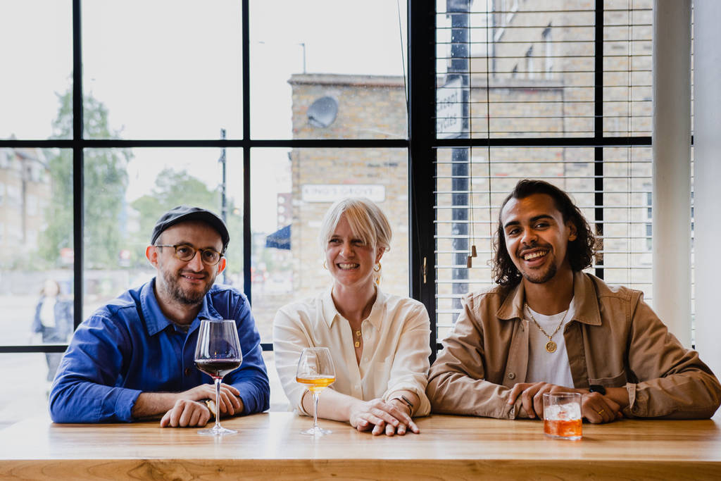 three people sat at a counter smiling at the camera, with two glasses of wine, and one cocktail, in front of them