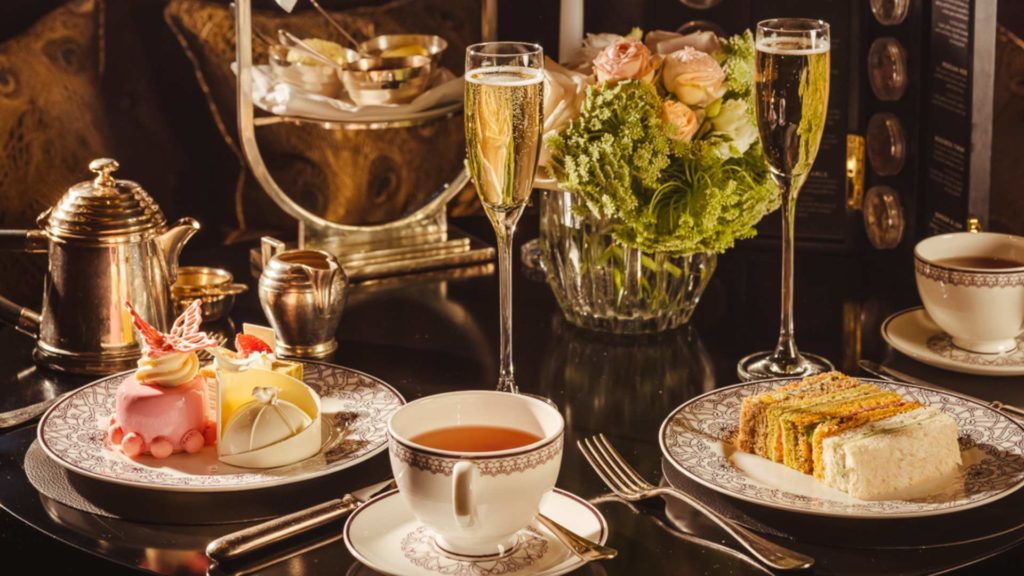 an afternoon tea spread from L'Oscar London, with a flute of champagne behind a cup of tea, a plate of sandwiches on the right, and a plate of desserts on the left