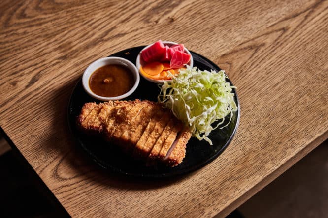 A plate of pork tonkatsu with cabbage, pickles, and brown sauce at Mu