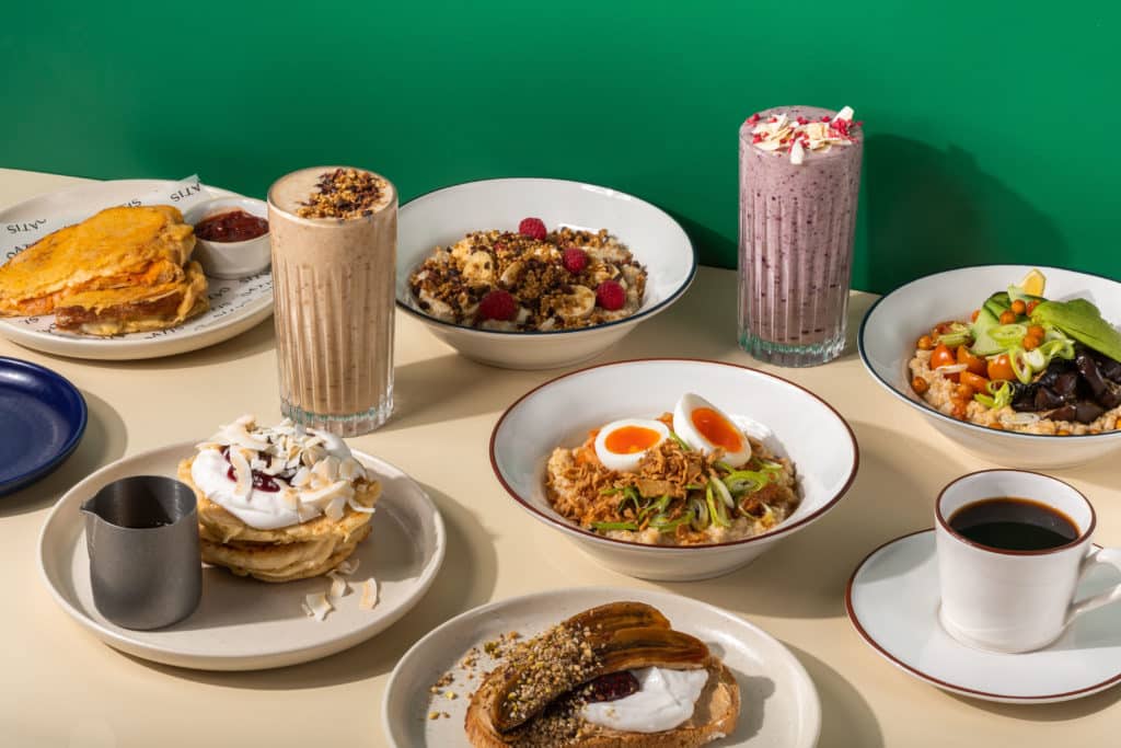 an array of oat bowls, pancakes, smoothies, and coffee placed on a table in front of a green wall