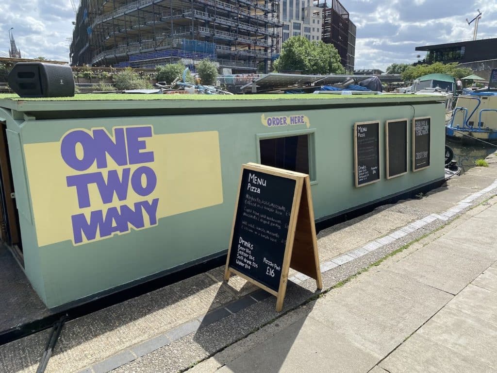 the 'one two many' pizza barge awaiting customers, with a menu board on the pavement next to its spot