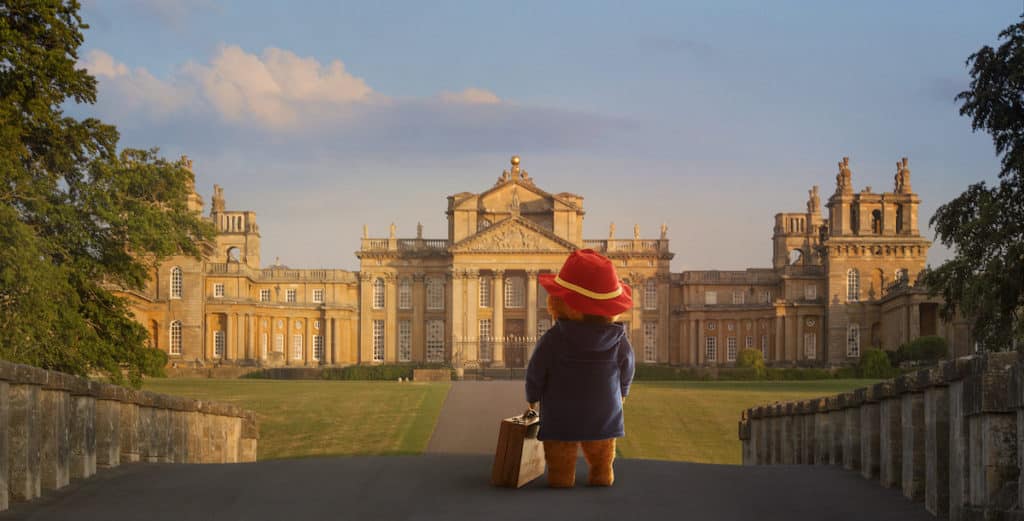 paddington bear, with his red hat and suit case, stands on a bridge look towards the regal Blenheim Palace