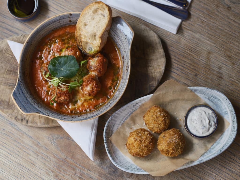 two plates of food, one containing meatballs in tomato sauce, and one with arancini balls