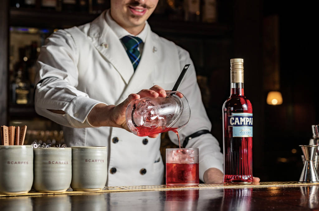 a bartender pouring a rich red coloured cocktail into a glass next to a bottle of campari