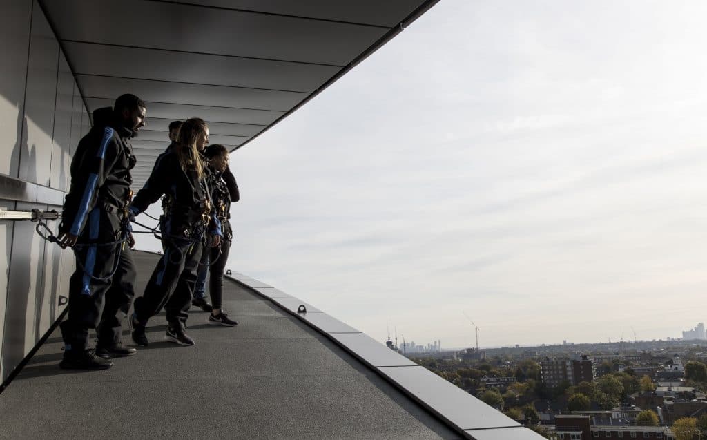 people stood, looking out at the view of London, from Tottenham stadium's The Dare Skywalk experience, wearing jumpsuits and harnesses that have them secured to the wall